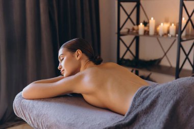 Rahatlamış müşteri, loş ışıklı bir spa odasında masaj terapisinin keyfini çıkarıyor. Mumlarla, sağlık ve vücut bakımını destekliyor.