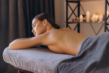 Masaj masasında uzanan genç bir kadın, arka planda mumlarla rahatlatıcı bir masaj terapisi seansının keyfini çıkarıyor ve huzurlu bir atmosfer yaratıyor.