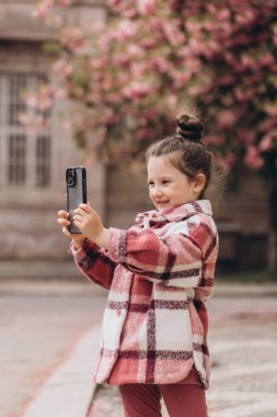 Kareli ceket giyen küçük bir kız bahar boyunca parkta akıllı telefonuyla selfie çekiyor.