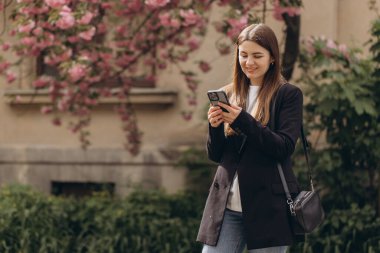 Genç yönetici, Sakura ağacının yanında yürürken akıllı telefondan sosyal medyada geziniyor.