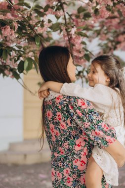 Sevgi dolu anne, gülümseyen kızını kiraz çiçeklerinin altında tutar, bir bahar bahçesinde hassas bir aile anını yakalar.