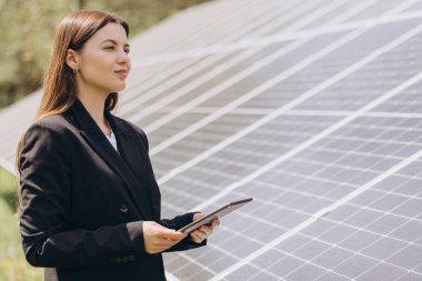 İş kadını elinde tablet tutuyor yenilenebilir enerji santralindeki güneş panellerini denetliyor, sürdürülebilir enerji üretimini kontrol ediyor