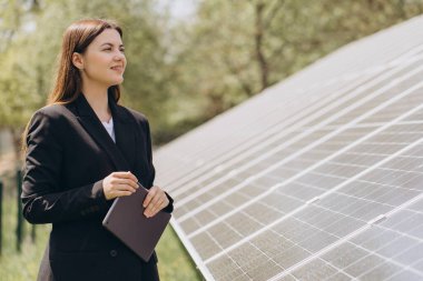İş kadını gülümsüyor ve elinde tabletle güneşli bir günde fotovoltaik enerji santralini inceliyor.