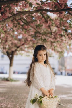 Çiçekli bir kiraz ağacının altında duran sevimli çocuk elinde bir buket taze çiçekle saman sepetini tutuyor.