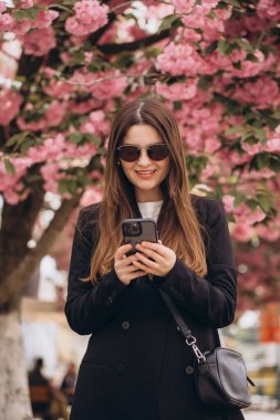 Baharda çiçek açan güzel sakura ağacının altında dururken cep telefonu kullanan genç bir iş kadını.