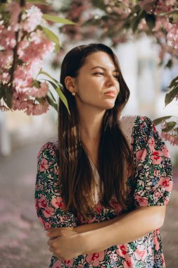 Uzun kahverengi saçlı, çiçek desenli elbisesi giyen güzel bir kadın. Çiçek açan Sakura ağacının altında duruyor. Bahar mevsiminin tadını çıkarıyor.