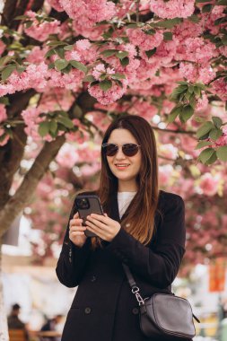 Genç bir iş kadını çiçek açan pembe sakura ağacının altında cep telefonuyla şehir parkında baharın özünü yakalıyor.