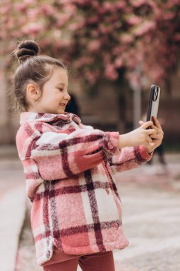 Neşeli bir bahar parkında güneşli bir günün tadını çıkaran gülümseyen anaokulu kızı akıllı telefonuyla mutlu bir şekilde selfie çekiyor.