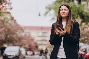 İş kadını, baharda ağaçlar ve park halindeki arabalarla sokakta yürüyen cep telefonu kullanıyor.