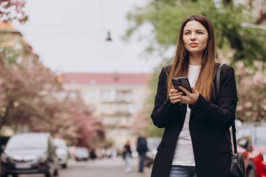 Genç yönetici cep telefonu uygulaması kullanıyor. Mesajlaşma mesajı. Baharda çiçek açan pembe ağaçlarla şehir caddesinde yürüyor.