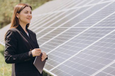 İş kadını elinde tablet tutarken ve gülümserken enerji santralindeki fotovoltaik güneş panellerini inceliyor