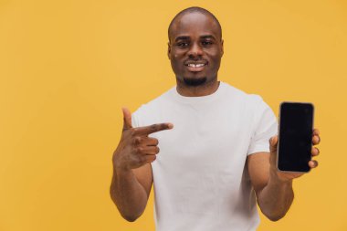 Gülümseyen Afro-Amerikan adam cep telefonuna boş ekranla işaret ediyor, cihazın modelini gösteriyor, uygulamayı öneriyor, sarı arka planda duruyor.