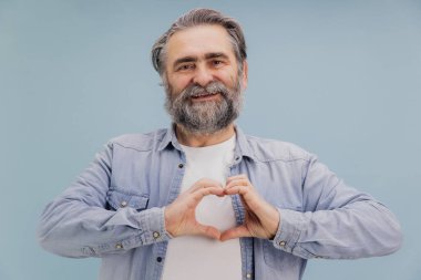 Gülümseyen sakallı son sınıf öğrencisi elleri ile canlı, mavi bir stüdyo geçmişine karşı kalp şekli oluşturuyor, sıcaklık ve şefkat saçıyor.