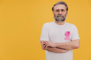 Göğüs kanseri farkındalığını destekleyen pembe kurdele takan kollarını kavuşturan kendinden emin bir kıdemli adamın portresi sarı arka planda duruyor.