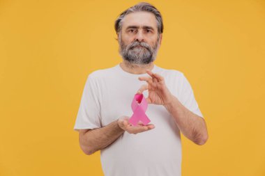 Sakallı son sınıf öğrencisi pembe kurdele gösteriyor, göğüs kanseri farkındalığını destekliyor, sarı arka planda.