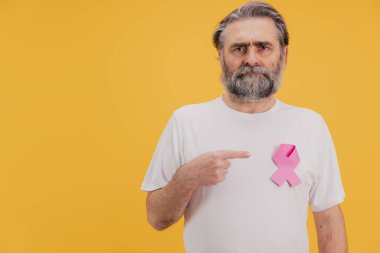 Ciddi kıdemli bir adam beyaz tişörtüne takılı pembe kurdeleyi göstererek meme kanseri farkındalığını arttırıyor.