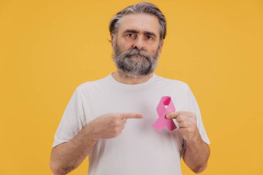Göğsüne takılı pembe kurdeleyi gösteren beyaz tişörtlü son sınıf sakallı adam göğüs kanseri farkındalığını destekliyor, sarı arka planda duruyor.