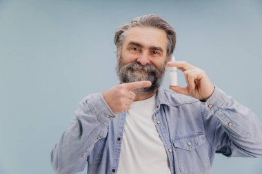 Soğuk algınlığı için burun spreyi tutan sakallı yaşlı bir adam parmağıyla işaret ediyor, açık mavi bir arka planda izole edilmiş.