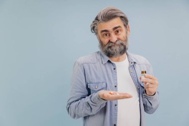 Elinde bir avuç dolusu renkli hap ve ilaç şişesiyle kameraya şüpheli bir ifadeyle bakan olgun bir adamın portresi.