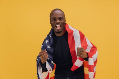 Heyecanlı Afrikalı Amerikalı adam, Amerikan bayrağına sarılmış, neşeli ve gururlu bir şekilde, canlı sarı bir arka plana karşı kutlama yapıyor.
