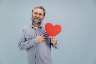 Gülümseyen sakallı, kot gömlekli, büyük kırmızı bir kalp taşıyan, sevgisini ve özenini gösteren, mavi stüdyo arka planında duran yaşlı adam.