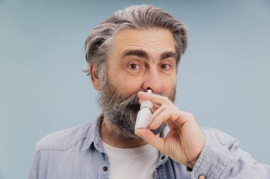 Gri saçlı ve sakallı olgun bir adam burun spreyi kullanıyor. Soğuk algınlığı ya da grip gibi bir hastalığın yol açtığı tıkanıklıktan kurtuluyor.