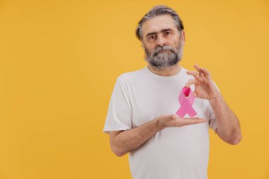 Sarı arka planda meme kanserini destekleyen pembe bir kurdele tutan sakallı yaşlı adam.