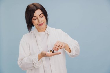 Orta yaşlı bir kadın eline şişeden hap döküyor, gülümsüyor, açık mavi bir arka planda.