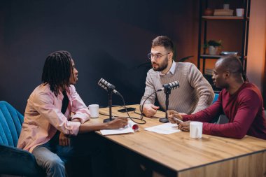 Professional radio hosts discussing podcast topic, interviewing guest while recording audio content in soundproof studio setting clipart