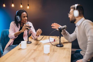 Erkekler podcast sırasında konuşuyor, profesyonel stüdyo ortamında mikrofonlar ve ses ekipmanları ile işbirliği içinde sohbet ediyorlar.