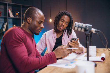 İki Afrikalı Amerikalı podcaster yeni bir bölüm kaydediyor ve bir akıllı telefona danışıyor, mikrofon ve belgelerle dolu bir masada oturuyor.