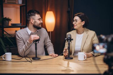 İki podcaster profesyonel bir stüdyoda bir bölüm kaydediyor. Sohbet ediyorlar ve görüşlerini mikrofonlar ve bir kamerayla paylaşıyorlar.