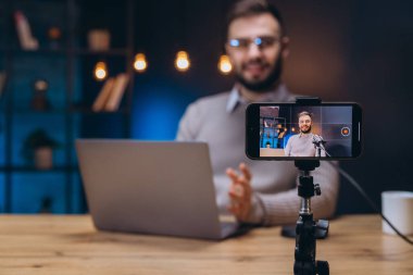 Podcaster mikrofona konuşuyor, akıllı telefon kaydı, stüdyo seansı sırasında yakınlarda dizüstü bilgisayar.