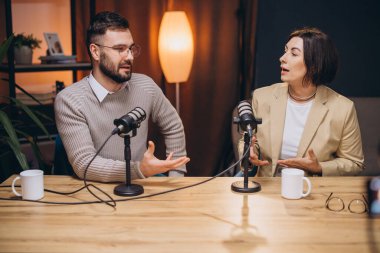 İki podcaster, bir erkek ve bir kadın, profesyonel bir stüdyoda bir bölüm kaydediyor, sohbet ediyorlar ve görüşlerini mikrofonlarla paylaşıyorlar.