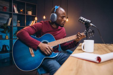Küçük ev stüdyosunda müzik kaydederken mavi akustik gitar çalan Afrikalı Amerikalı müzisyen.