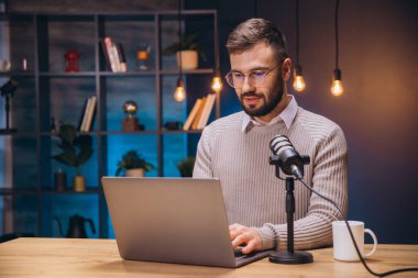 Erkek podcaster ev kayıt kurulumundan dizüstü bilgisayarda çalışırken yoğunlaştırıcı mikrofona konuşuyor