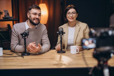 İki podcaster, mikrofon ve kamerayla çevrili modern bir stüdyoda, dinamik konuşmaları kaydeden bir video yayını yapıyorlar.
