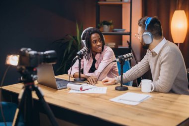 Erkek podcaster 'lar canlı radyo programı kaydediyor. Profesyonel mikrofonlarla ses panosunda oturuyorlar ve kulaklık takıyorlar.
