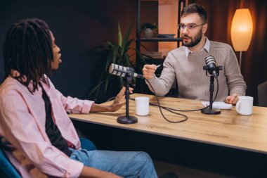 Genç podcaster 'lar birlikte ses kaydı yapıyorlar, kulaklık takıyorlar ve ses geçirmez stüdyo ortamında profesyonel mikrofonlara konuşuyorlar.