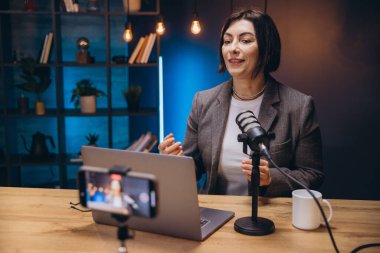 Profesyonel bir mikrofon ve dizüstü bilgisayar kullanarak ev stüdyosundan podcast bölümü kaydederken orta yaşlı bir kadın etkili jest yapıyor.