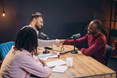 İki radyo sunucusu, profesyonel bir stüdyoda konuklarıyla podcast yaparken el sıkışıyor.