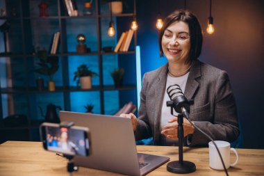 Neşeli kadın ses içeriği yaratıyor, mikrofona konuşuyor ve ev stüdyosunda dizüstü bilgisayar ve akıllı telefon kullanarak podcast yapıyor.