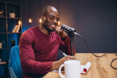 Genç Afrikalı Amerikalı adam sesini bir podcast bölümü için kaydediyor, mikrofonla konuşuyor ve notlar alıyor.