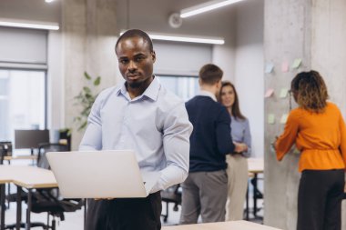 Laptop kullanan Afrikalı Amerikalı bir adam, modern ofis ortamında işbirliği yapan profesyoneller, iş ortamında çeşitliliği ve takım çalışmasını vurgulayan