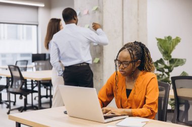 Genç siyahi girişimci şirket ofisinde dizüstü bilgisayar kullanıyor, arka plandaki projede iş birliği yaparken işine konsantre oluyor