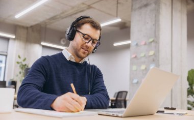 Bir ofis çalışanı, notlar alırken ve dizüstü bilgisayar kullanırken çevrimiçi bir toplantıya katılıyor ve teknolojiyle dolu modern bir çalışma alanını sergiliyor.