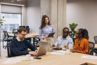 Modern, açık konsept ofis ortamında dijital cihazlar kullanırken, çoklu etnik gruplar işbirliği yapıyor, görüşleri paylaşıyor