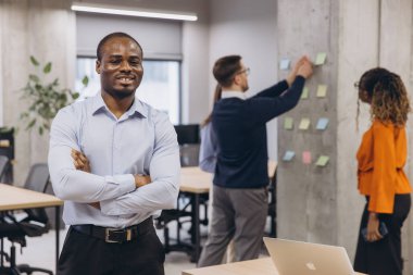 Gülümseyen Afrikalı Amerikalı erkek, profesyonel takım işbirliği yapıyor, beton duvarın yanında beyin fırtınası stratejisi uyguluyor, modern çalışma alanında renkli yapışkan notlar düzenliyor.