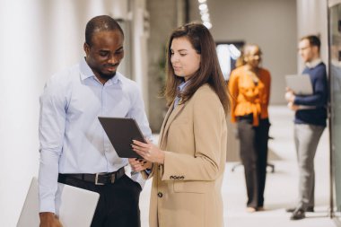 Bir ofis koridorunda iş birliği yapan iş arkadaşları, etkin iletişim ve planlama için dijital tablet kullanırken iş hakkında tartışıyorlar.