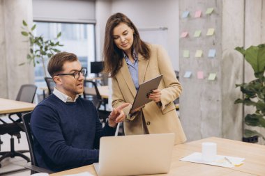 Profesyonel ekip işbirliği yapıyor, yeşil bitkiler ve çağdaş çalışma alanındaki renkli yapışkan notlarla çevrili dizüstü bilgisayar ve tablet üzerinde çalışıyor.